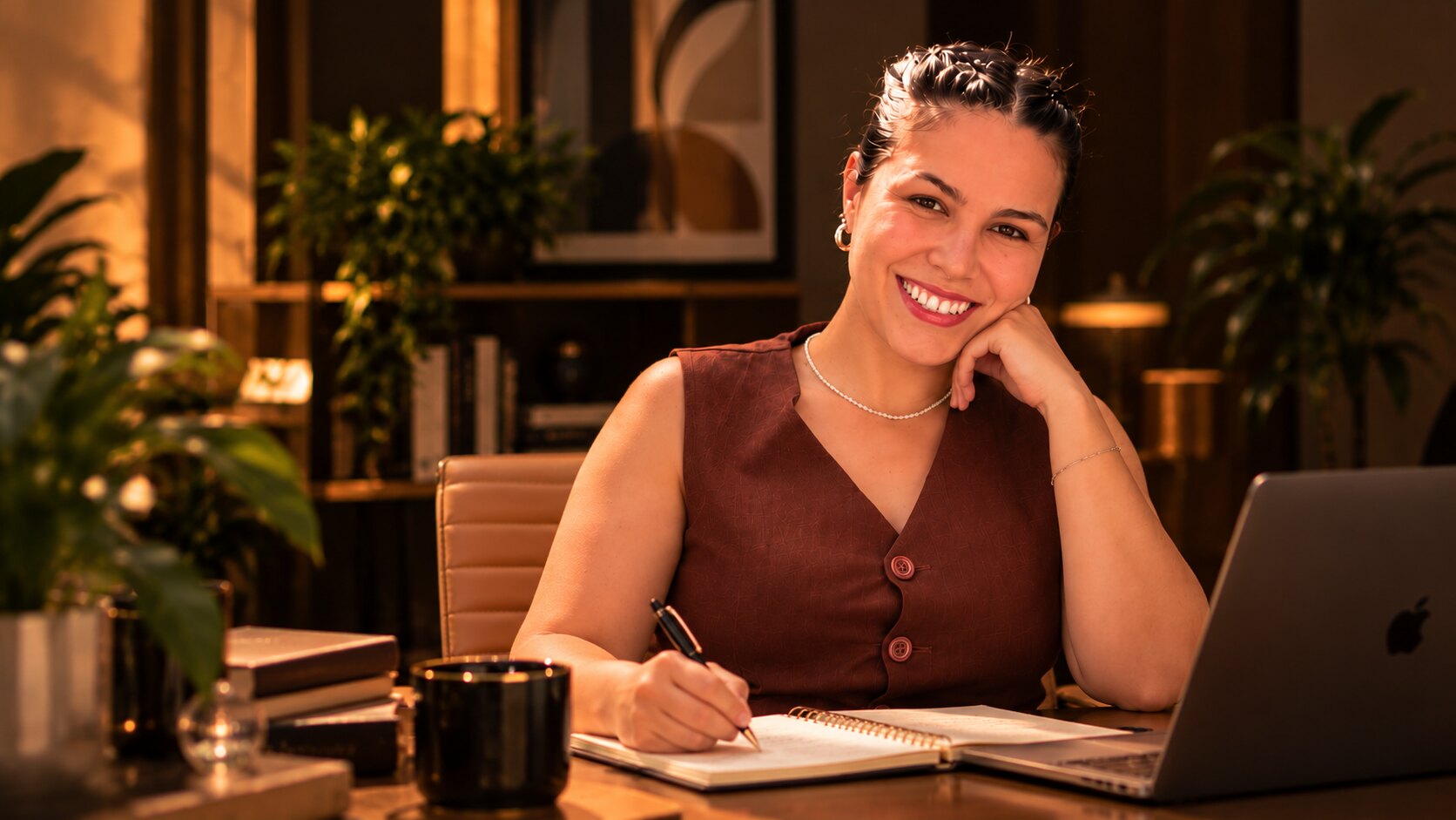 Ayla Camargo sorrindo, sentada à mesa do escritório, escrevendo em um caderno